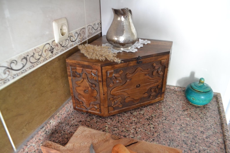May include: A wooden chest with carved floral designs, a silver pitcher, and a small turquoise jar with a lid. The chest is brown, and the pitcher has a hammered texture. A sprig of dried grass sits on top of the chest.
