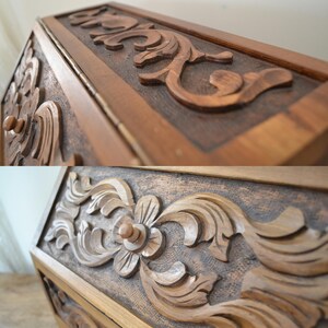 May include: A wooden box featuring intricate hand-carved floral and scrolling leaf patterns on its exterior panels. The wood has a medium brown finish with a textured, darkened background behind the raised, smooth-carved decorative elements.