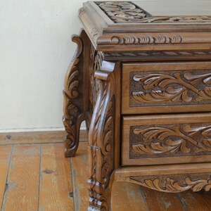Puede incluir: Una mesa auxiliar de madera con dos cajones, ambos con intrincados dise&ntilde;os florales tallados. La mesa tiene patas ornamentadas con detalles tallados y un acabado marr&oacute;n oscuro.