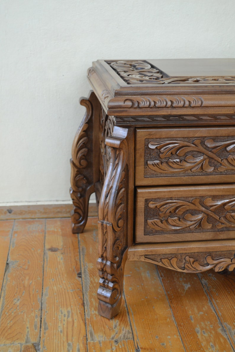 Puede incluir: Una mesa auxiliar de madera con dos cajones, ambos con intrincados dise&ntilde;os florales tallados. La mesa tiene patas ornamentadas con detalles tallados y un acabado marr&oacute;n oscuro.