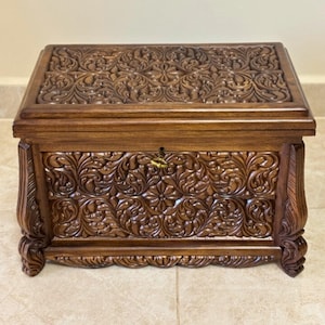Puede incluir: Un cofre de madera marrón oscuro con intrincadas tallas florales en la tapa, el frente y los lados. El cofre tiene forma rectangular, una cerradura y patas ornamentadas. La madera tiene un acabado pulido.