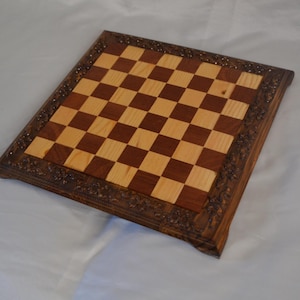 K&ouml;nnte beinhalten: Ein Schachbrett aus Holz mit einem dunkelbraunen Rahmen und einer hellen Holzspielfl&auml;che. Der Rahmen hat ein geschnitztes Design.