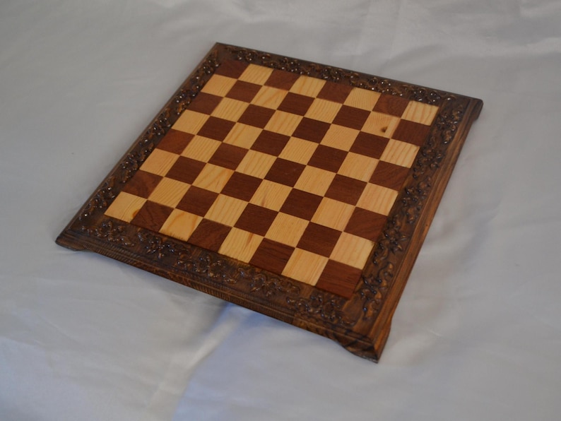 K&ouml;nnte beinhalten: Ein Schachbrett aus Holz mit einem dunkelbraunen Rahmen und einer hellen Holzspielfl&auml;che. Der Rahmen hat ein geschnitztes Design.