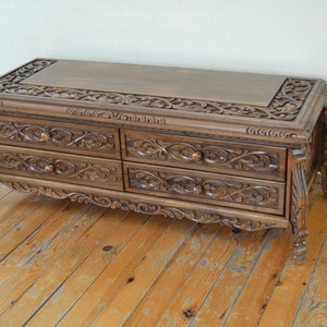 Puede incluir: Una mesa de centro de madera con cuatro cajones y tallas intrincadas. La mesa tiene un acabado marr&oacute;n oscuro y patas ornamentadas.