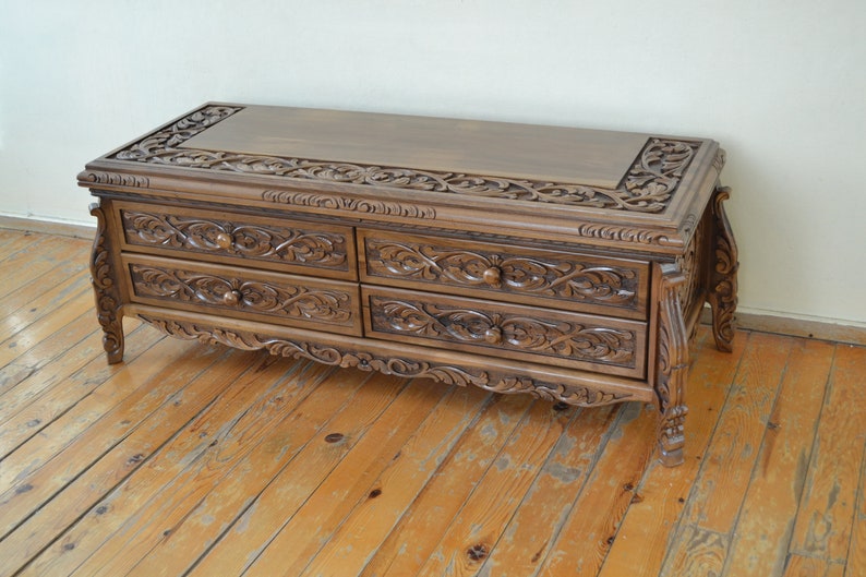 Puede incluir: Una mesa de centro de madera con cuatro cajones y tallas intrincadas. La mesa tiene un acabado marr&oacute;n oscuro y patas ornamentadas.