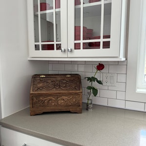 May include: A dark brown wooden bread bin featuring intricate floral carvings on the front panel and lid, equipped with gold-toned metal hinges. Beside the bread bin stands a single red rose in a clear glass vase with a decorative silver base.