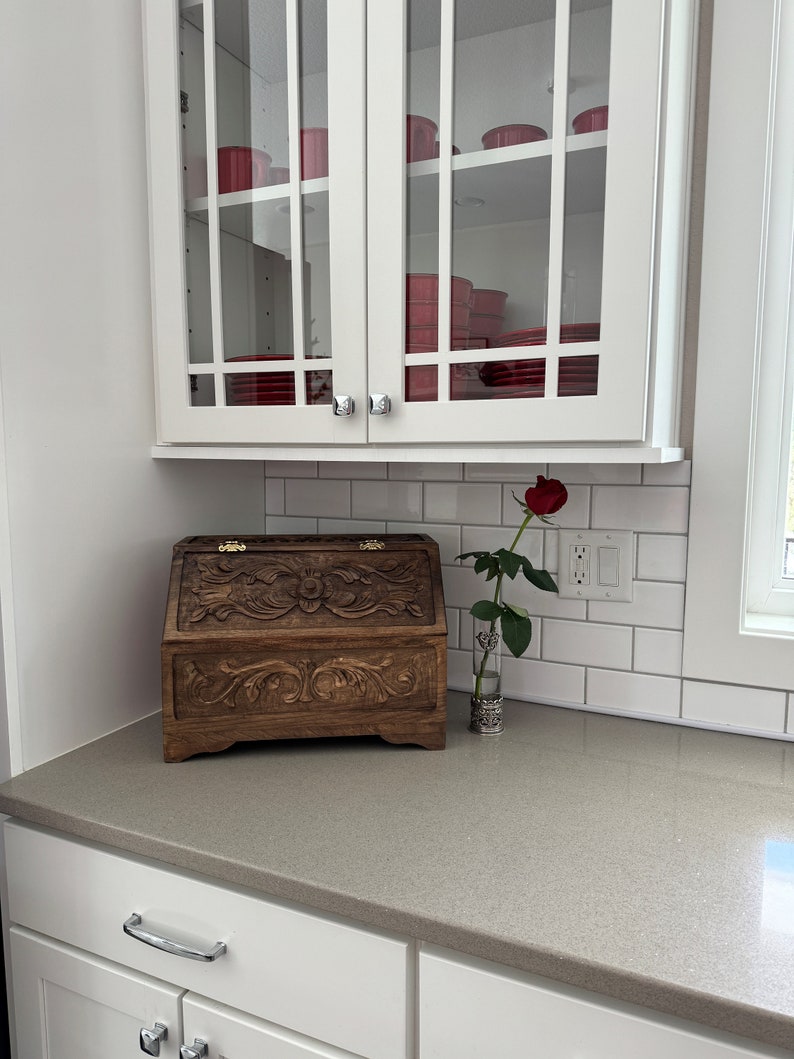 May include: A dark brown wooden bread bin featuring intricate floral carvings on the front panel and lid, equipped with gold-toned metal hinges. Beside the bread bin stands a single red rose in a clear glass vase with a decorative silver base.