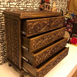 Puede incluir: Una c&oacute;moda de madera con cinco cajones, cada uno tallado intrincadamente con motivos florales. La c&oacute;moda es de un color marr&oacute;n intenso y tiene un acabado ligeramente desgastado.