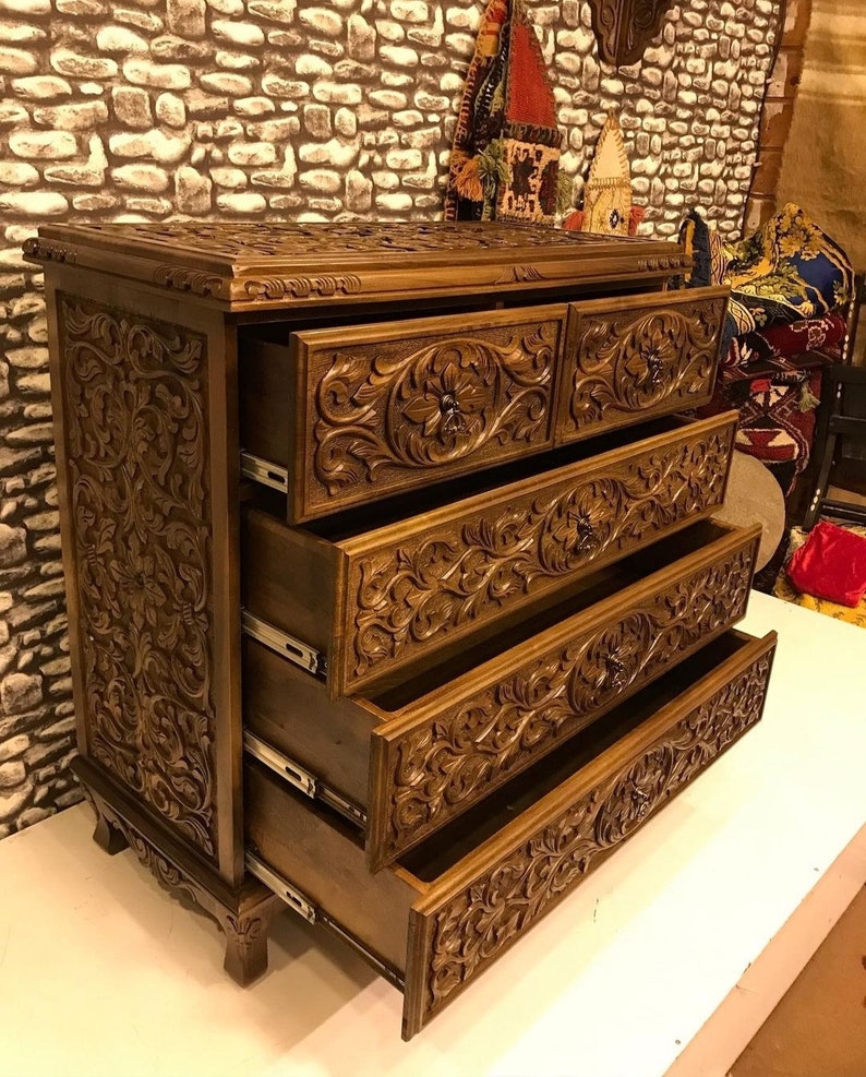 Puede incluir: Una c&oacute;moda de madera con cinco cajones, cada uno tallado intrincadamente con motivos florales. La c&oacute;moda es de un color marr&oacute;n intenso y tiene un acabado ligeramente desgastado.