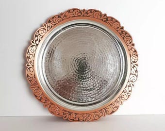 May include: A round metal tray with a hammered silver center and a copper-colored, ornate, scalloped edge. The tray has a decorative floral pattern around the rim. The tray is likely used for serving or display.