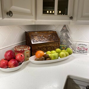May include: A white bowl filled with four red pomegranates, a decorative tin, a wooden bread box with floral carvings, and an oval white platter holding an orange, several kiwis, and green pears, plus a decorative tree and a sign reading I JUST WANT TO BAKE STUFF AND WATCH CHRISTMAS MOVIES.