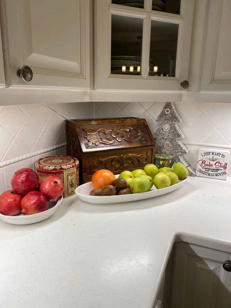 May include: A white bowl filled with four red pomegranates, a decorative tin, a wooden bread box with floral carvings, and an oval white platter holding an orange, several kiwis, and green pears, plus a decorative tree and a sign reading I JUST WANT TO BAKE STUFF AND WATCH CHRISTMAS MOVIES.
