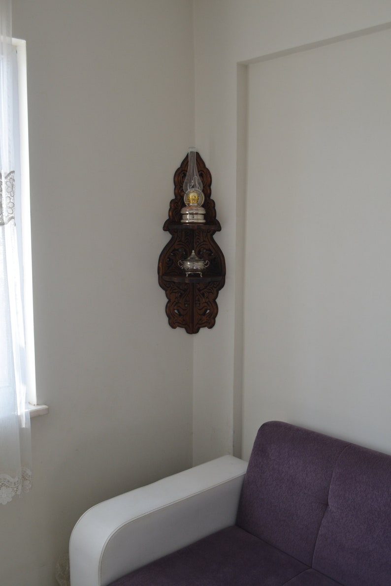 May include: A wooden corner shelf with intricate carvings. The shelf has two tiers and features a glass oil lamp with a silver base and a small silver bowl.