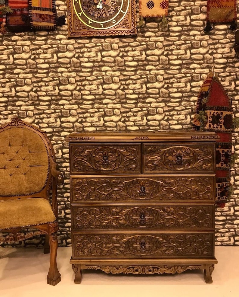 Puede incluir: Una c&oacute;moda de madera con cinco cajones, cada uno tallado intrincadamente con motivos florales. La c&oacute;moda es de un color marr&oacute;n intenso y tiene un acabado ligeramente desgastado. La c&oacute;moda est&aacute; colocada sobre un fondo de pared de piedra.