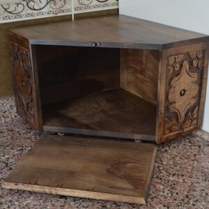 May include: A dark brown, corner cabinet with a hinged door. The cabinet has carved floral designs on the sides and a triangular shape. The door is open, revealing the interior.