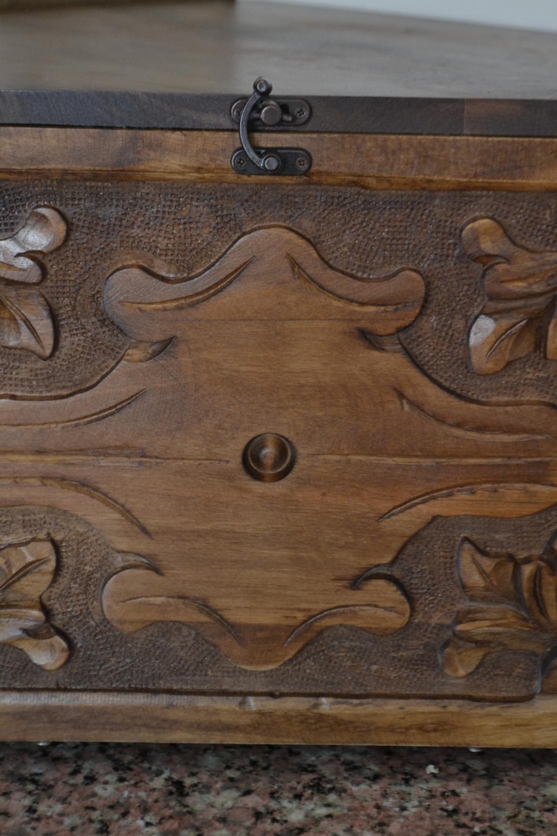 May include: A wooden chest with intricate carved floral and scroll designs. The chest is stained a warm brown color and features a metal clasp. The carved details are highlighted with a darker shade, adding depth to the design.