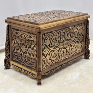 Puede incluir: Un cofre de madera con intrincadas tallas florales. El cofre rectangular tiene una tapa con bisagras y descansa sobre patas cortas. La madera tiene un tono marrón cálido, y las tallas ornamentadas crean un mueble de almacenamiento decorativo.