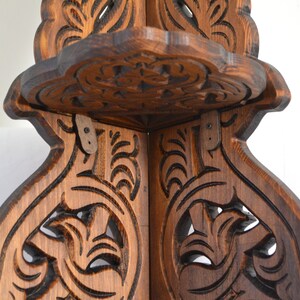 May include: A wooden corner shelf with intricate carvings. The shelf has a dark brown stain and is made of solid wood. The shelf is designed to fit in a corner and has two shelves.