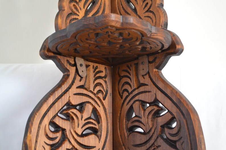 May include: A wooden corner shelf with intricate carvings. The shelf has a dark brown stain and is made of solid wood. The shelf is designed to fit in a corner and has two shelves.