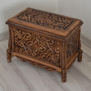Puede incluir: Un cofre de madera con intrincados tallados en un patrón floral. El cofre tiene una tapa con bisagras y se asienta sobre cuatro patas.
