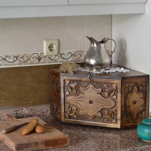 May include: A wooden bread box with carved floral designs, a silver pitcher, and a small turquoise jar with a lid sit on a countertop. A wooden cutting board with a knife and bread is in the foreground.