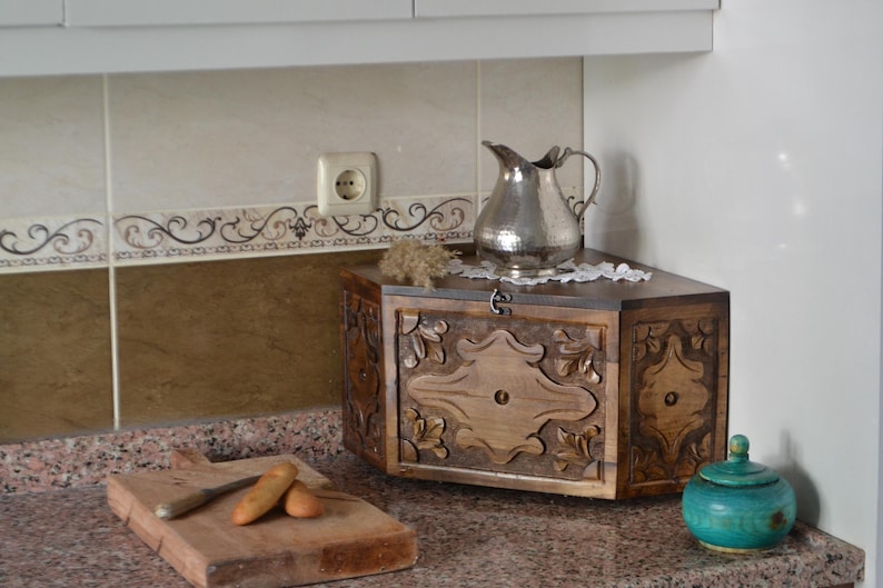 May include: A wooden bread box with carved floral designs, a silver pitcher, and a small turquoise jar with a lid sit on a countertop. A wooden cutting board with a knife and bread is in the foreground.