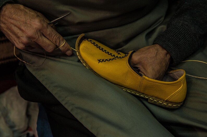 Puede incluir: Una persona est&aacute; cosiendo a mano un zapato de cuero amarillo con costuras negras. El zapato tiene una punta puntiaguda y un dise&ntilde;o simple.
