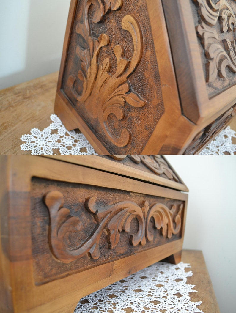 May include: A brown wooden box featuring intricate relief carvings of scrolling acanthus leaves and floral motifs. The surface displays a textured, stippled background behind the raised carvings, with a smooth frame border and small feet.