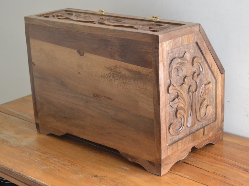 May include: A rectangular wooden bread bin crafted from dark brown wood with visible grain. The side panel features an intricate carved floral relief pattern. The top lid is hinged with gold-coloured metal hardware and also displays a carved design.