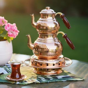 Könnte beinhalten: Ein kupfernes Teeservice mit gehämmertem Muster, bestehend aus einer zweistöckigen Teekanne mit Holzgriffen. Das Set beinhaltet einen Warmhalteständer und ein kleines Glas Tee auf einer Untertasse, auf einem Tisch mit Blumenarrangement.