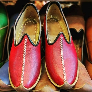 Puede incluir: Dos zapatos de cuero rojo con ribetes negros y costuras blancas. Los zapatos tienen una punta puntiaguda y una parte trasera cerrada.