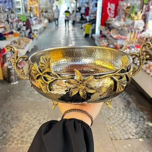 May include: A hammered copper bowl with ornate gold leaf designs. The bowl has three legs and two handles. The bowl is ideal for serving snacks or fruit.