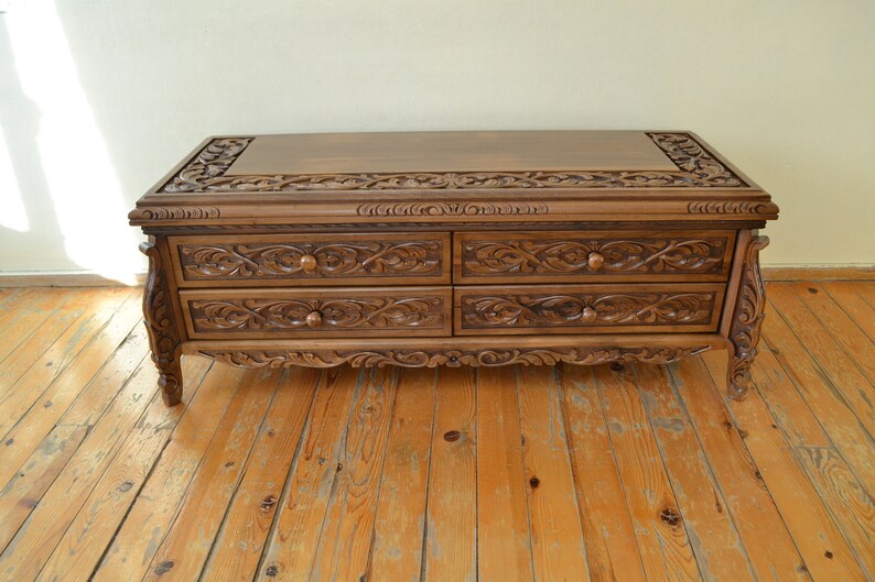 Puede incluir: Una mesa de centro de madera marr&oacute;n oscuro con cuatro cajones. Los cajones est&aacute;n tallados con intrincados dise&ntilde;os florales. La mesa tiene una tapa rectangular y se apoya en cuatro patas.
