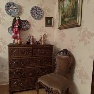 Puede incluir: Una c&oacute;moda de madera con seis cajones, tallas intrincadas y un acabado marr&oacute;n. Una silla de terciopelo marr&oacute;n con tallas ornamentadas se encuentra junto a la c&oacute;moda. La pared detr&aacute;s de la c&oacute;moda est&aacute; cubierta con papel tapiz floral con un patr&oacute;n rosa y blanco.