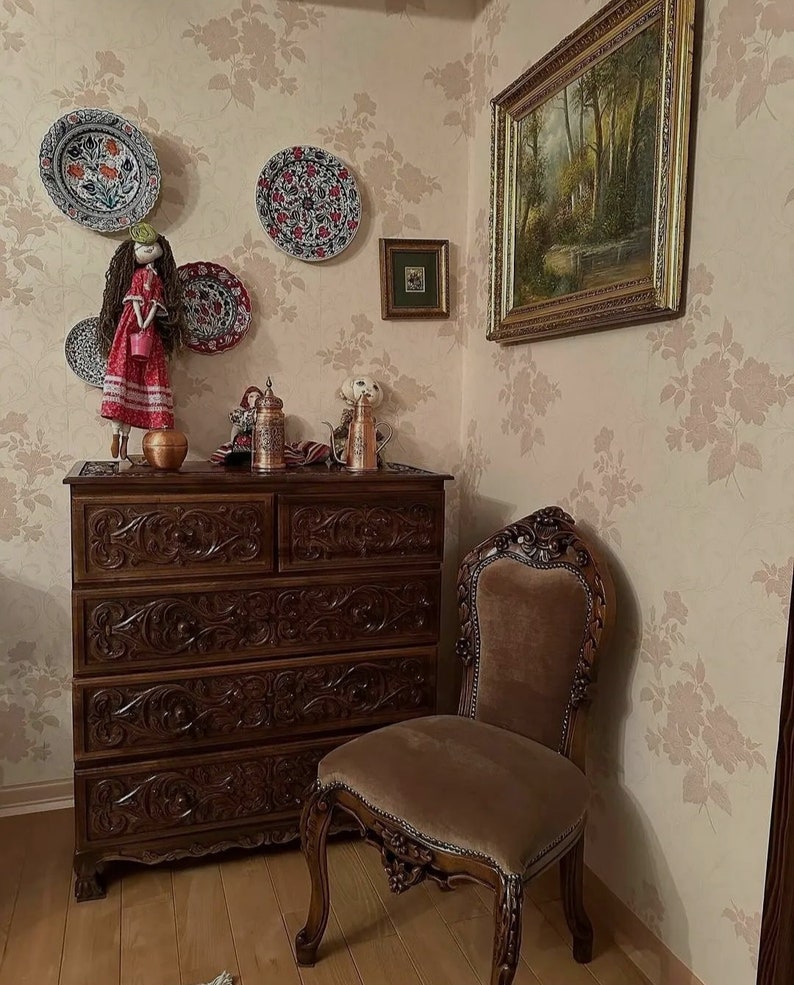 Puede incluir: Una c&oacute;moda de madera con seis cajones, tallas intrincadas y un acabado marr&oacute;n. Una silla de terciopelo marr&oacute;n con tallas ornamentadas se encuentra junto a la c&oacute;moda. La pared detr&aacute;s de la c&oacute;moda est&aacute; cubierta con papel tapiz floral con un patr&oacute;n rosa y blanco.