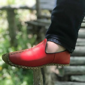 Puede incluir: Un mocas&iacute;n de cuero rojo con un dise&ntilde;o cosido y una suela marr&oacute;n. El mocas&iacute;n lo lleva puesto una persona que lleva pantalones vaqueros azules.