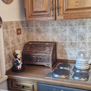 May include: A dark brown wooden bread bin with intricate floral carvings on the front panel, featuring a hinged lid, positioned on a kitchen counter next to a chef figurine and a white kettle on a four-burner electric stove top.