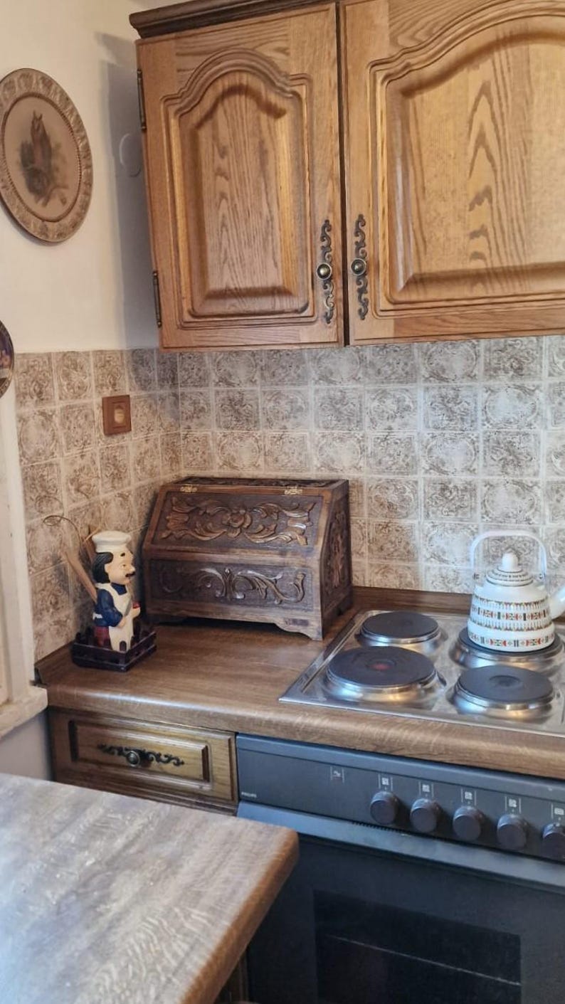 May include: A dark brown wooden bread bin with intricate floral carvings on the front panel, featuring a hinged lid, positioned on a kitchen counter next to a chef figurine and a white kettle on a four-burner electric stove top.