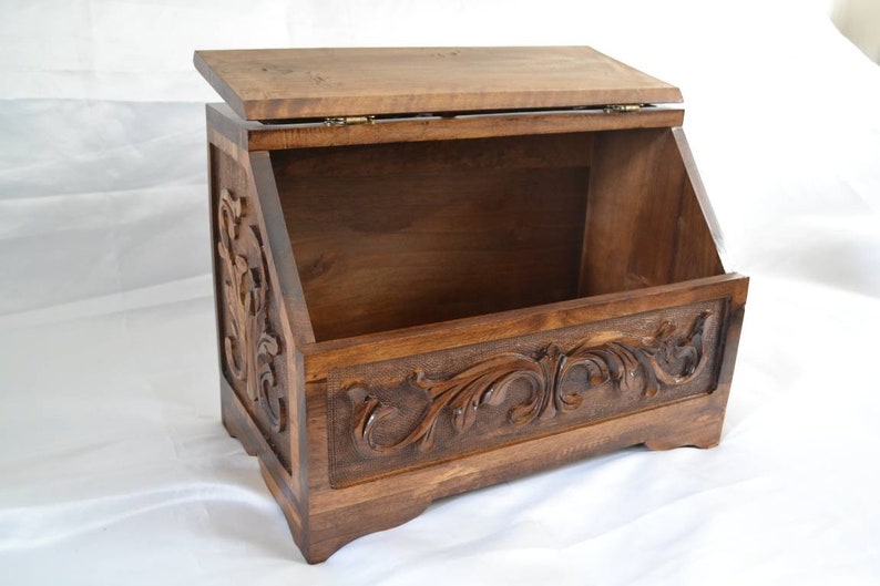 May include: A dark brown wooden storage bin with a hinged lid and an open front. The exterior features intricate carved scrollwork patterns on the front and side panels. The box has a rectangular base with four small feet and a smooth finish.