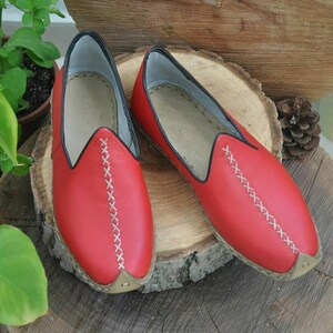 Puede incluir: Un par de zapatillas de cuero rojo con costuras blancas. Las zapatillas tienen una punta puntiaguda y una suela plana.