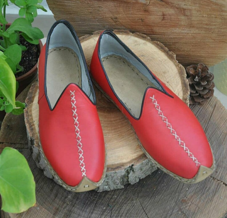 Puede incluir: Un par de zapatillas de cuero rojo con costuras blancas. Las zapatillas tienen una punta puntiaguda y una suela plana.