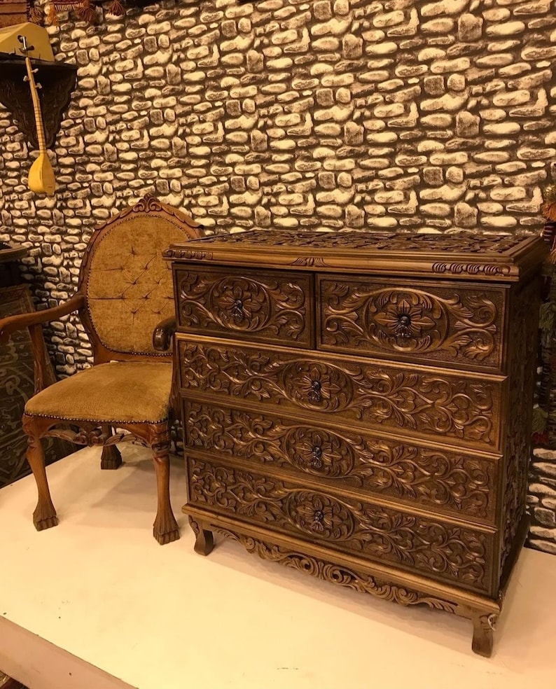 Puede incluir: Una c&oacute;moda de madera marr&oacute;n oscuro con cinco cajones, cada uno tallado intrincadamente con dise&ntilde;os florales. La c&oacute;moda tiene patas ornamentadas y una parte superior ligeramente curvada.