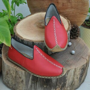Puede incluir: Un par de zapatos rojos de cuero sin cordones con costuras blancas. Los zapatos tienen una punta puntiaguda y una suela plana.
