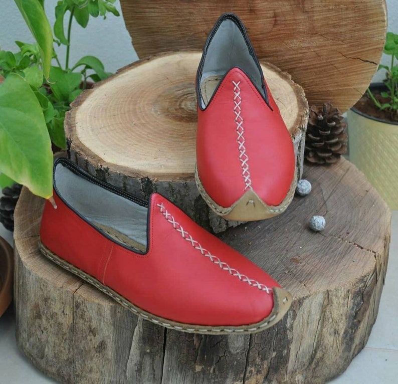 Puede incluir: Un par de zapatos rojos de cuero sin cordones con costuras blancas. Los zapatos tienen una punta puntiaguda y una suela plana.