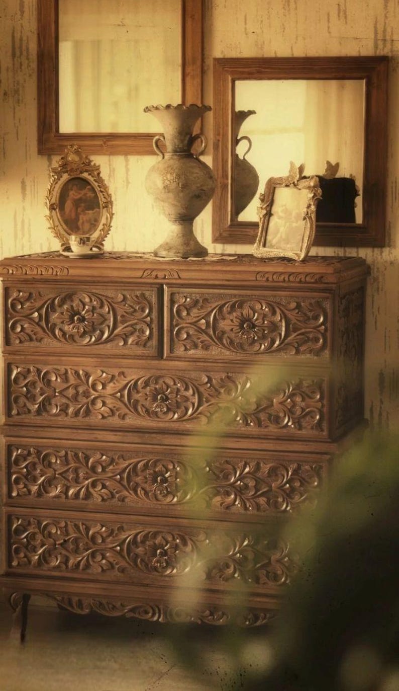 Puede incluir: Una c&oacute;moda de madera con intrincados tallados en forma de flores. La c&oacute;moda tiene seis cajones y se encuentra sobre un suelo de madera. Hay tres cuadros enmarcados y un jarr&oacute;n en la parte superior de la c&oacute;moda.
