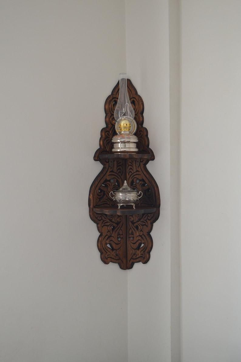 May include: A dark brown wooden corner shelf with intricate carvings. The shelf has two tiers and features a glass oil lamp and a small silver bowl.