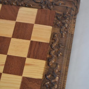 K&ouml;nnte beinhalten: Ein Schachbrett aus Holz mit einem geschnitzten, verzierten Rahmen. Das Brett hat ein Schachbrettmuster aus hellem und dunklem Holz.