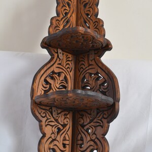 May include: A dark brown wooden corner shelf with intricate carvings. The shelf has three tiers and is designed to fit in a corner.