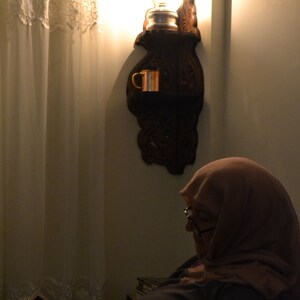 May include: A woman wearing a headscarf sits in a chair and reads a book. A wooden wall sconce with a light bulb and a copper mug is on the wall behind her.