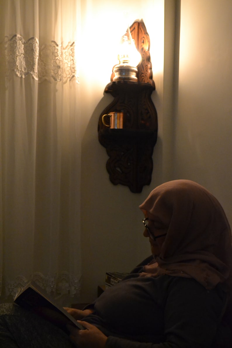 May include: A woman wearing a headscarf sits in a chair and reads a book. A wooden wall sconce with a light bulb and a copper mug is on the wall behind her.